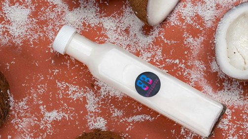 White coconut cocktail in a bottle next to coconuts sprinkles and fruits in the background