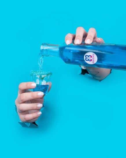 Two hands ripping through blue background pouring blue candy cocktail inside a shot glass