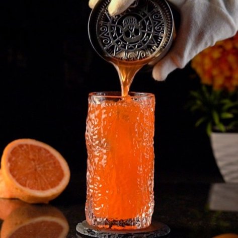 Orange cocktail being poured from skull Hawthorne strainer into possessed ancient glass