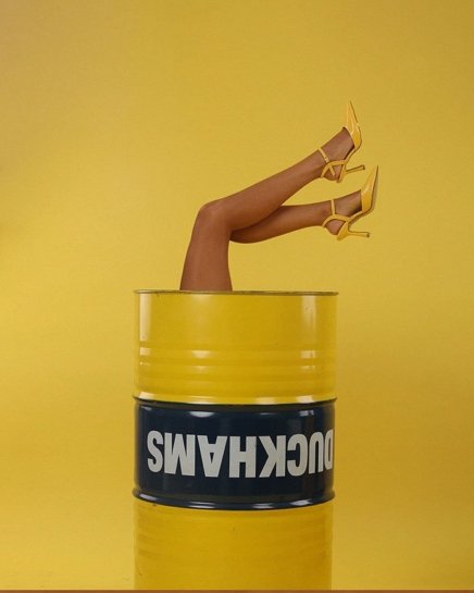 Woman model sitting inside a barrel with her feet upwards wearing a yellow pair of fashion heels