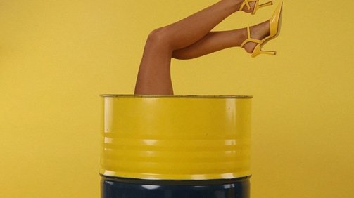 Woman model sitting inside a barrel with her feet upwards wearing a yellow pair of fashion heels